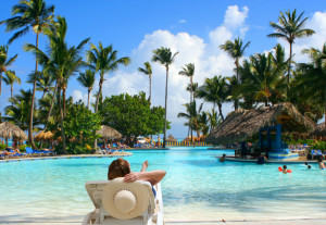 woman sitting poolside holding a straw beach hat background is a tropical pool bar with families enjoying their vacation